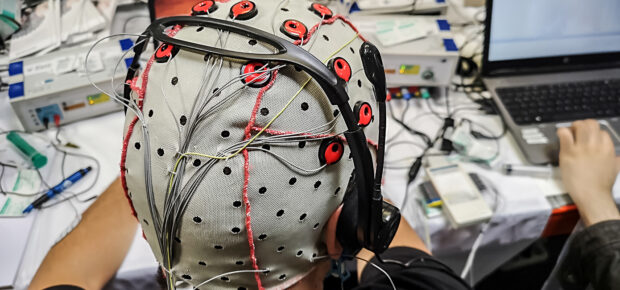 The electroencephalogram (EEG) head cap with flat metal discs (electrodes) in a science lab with laptops blurred at the background