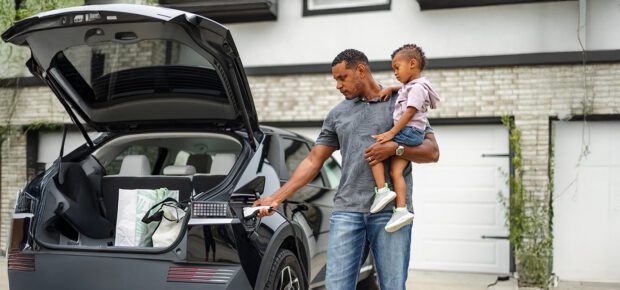 A father holds his young son while plugging an electric vehicle into the charging port at their home. The image highlights sustainable energy practices and reflects a modern, eco-friendly lifestyle.